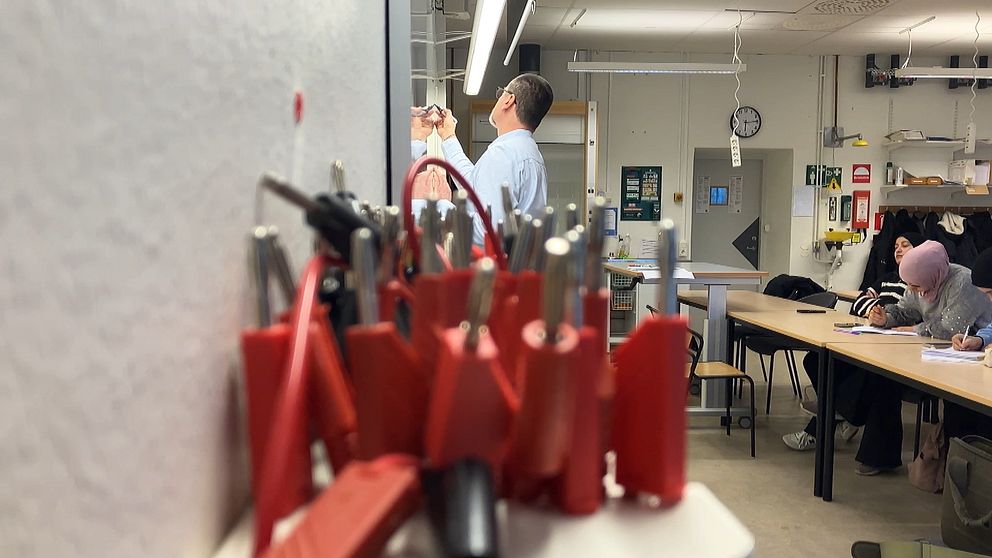 Ett klassrum fullt med elever och föreläsningsmaterial. I förgrunden syns kablar och elektronikverktyg, medan en man i bakgrunden skriver på en whiteboard.