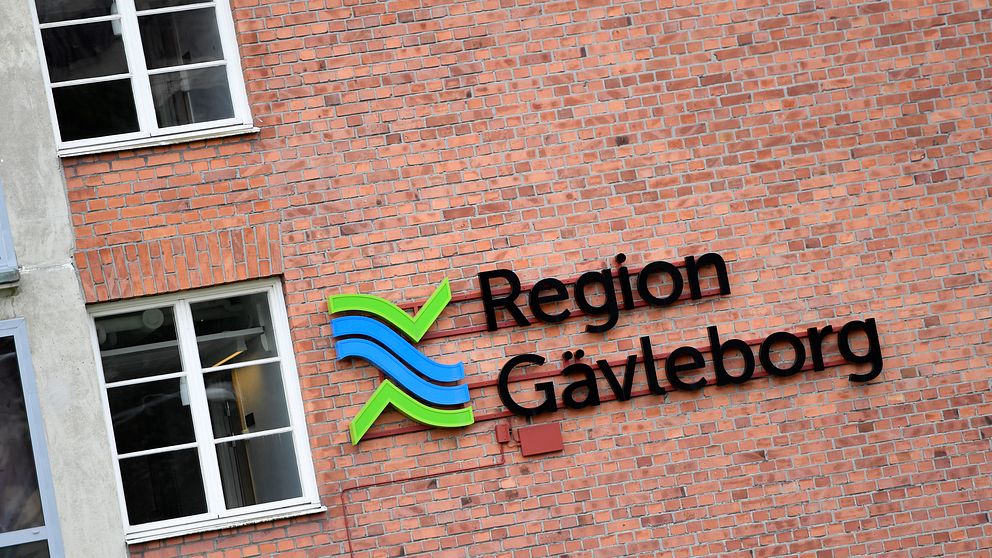 Logotypen för Region Gävleborg på en tegelvägg, med en grafisk design som inkluderar blå och gröna färger samt texten ”Region Gävleborg”. Flera fönster är synliga i närheten.
