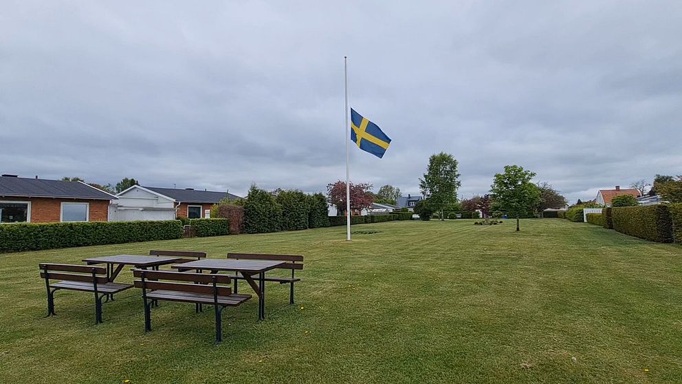Svensk flagga på halv stång i en park med tomma picknickbord under molnig himmel.