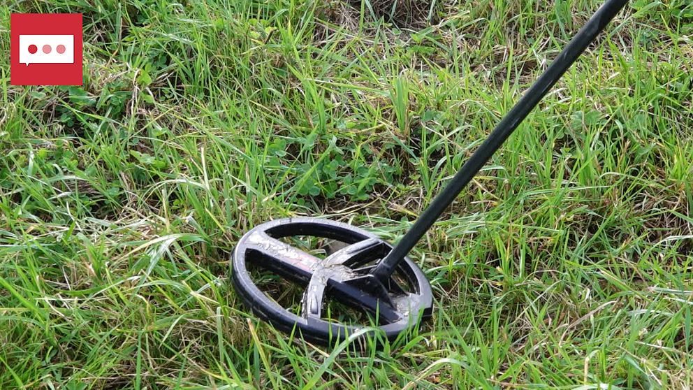 En metalldetektor som står i gräset, med fokus på den rundade sökspolen nära markytan. Gräset är frodigt och grönt runt detektorn.