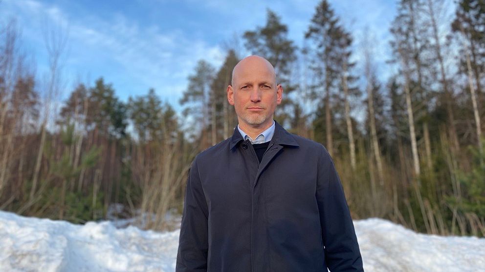 En person står framför en snötäckt mark med träd i bakgrunden under en klar himmel. Personen är klädd i en mörk jacka och skjorta, vilket ger en känsla av vinter och utomhusaktiviteter.