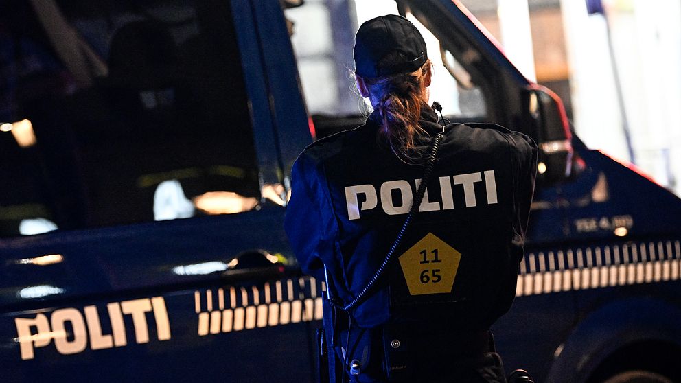 Polis i uniform står med ryggen mot kameran vid en blå polisiär bil, med ordet ”POLITI” synligt på både bilen och uniformen.