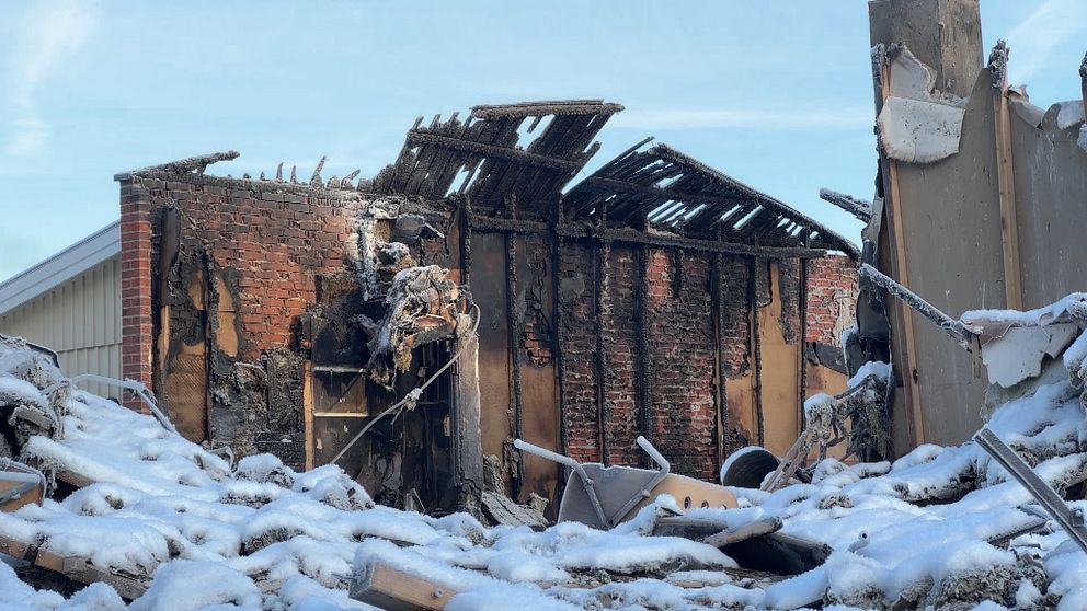 Resterna av en branddrabbad byggnad är synliga, där tegelväggar är blottade och taket har kollapsat. Snö täcker marken runtomkring, vilket tyder på kallt väder. Det finns skräp och rivningsmaterial spritt på marken, vilket visar att byggnaden har genomgått betydande skador.