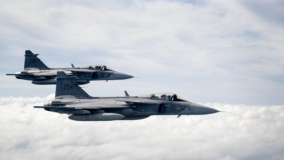 Två jetplan av typen JAS 39 Gripen flyger i formation över molniga himlar.