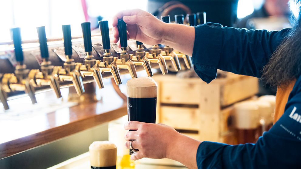 En person häller upp ett mörkt öl från en tappkran i en bar, med flera tappkranar synliga i bakgrunden. Ölet har ett stort, skummande huvud och det finns flera glas med öl på bardisken.