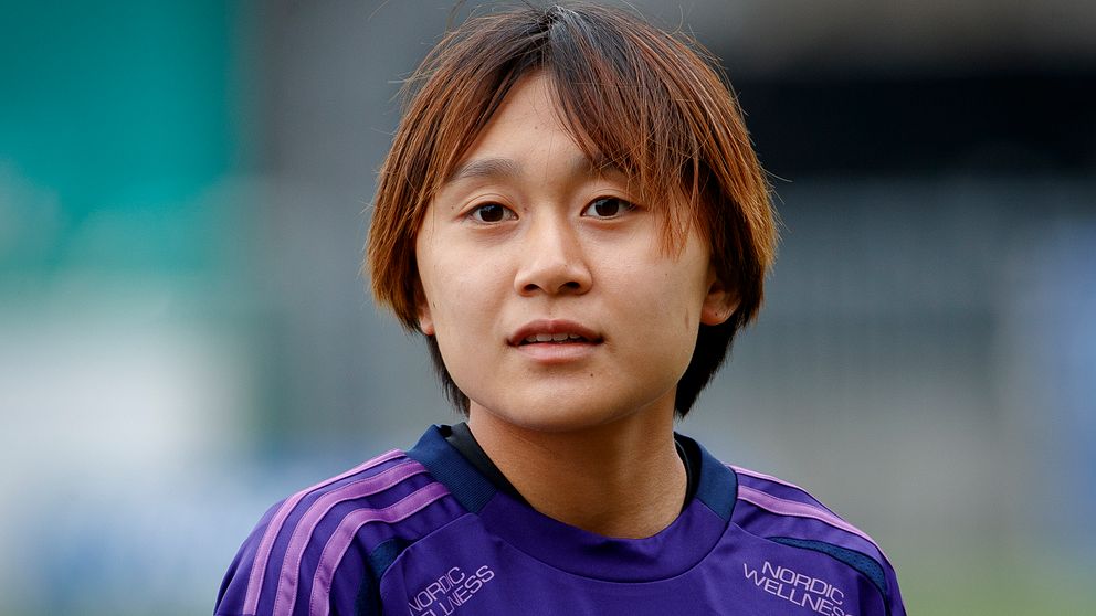 En kvinna i en lila sporttröja står framför en suddig bakgrund, troligtvis på en fotbollsplan. Hennes uppsyn är fokuserad, vilket antyder att hon är i en träningssituation eller förbereder sig för en match.