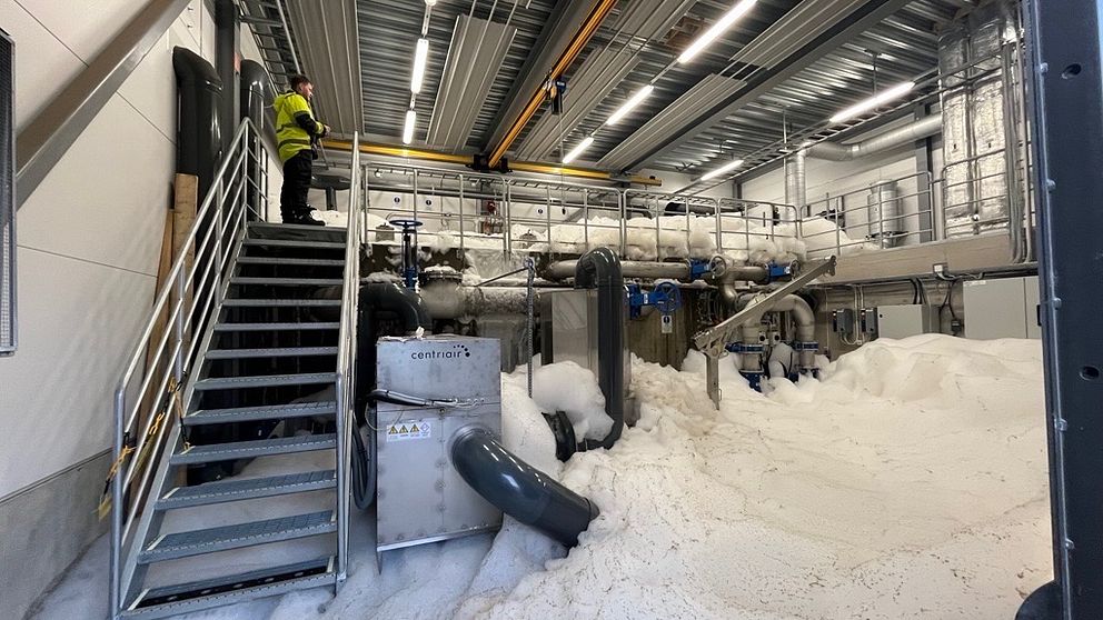 En person med skyddsutrustning står på en trappa bredvid stora mängder skum på golvet i en industriell anläggning.