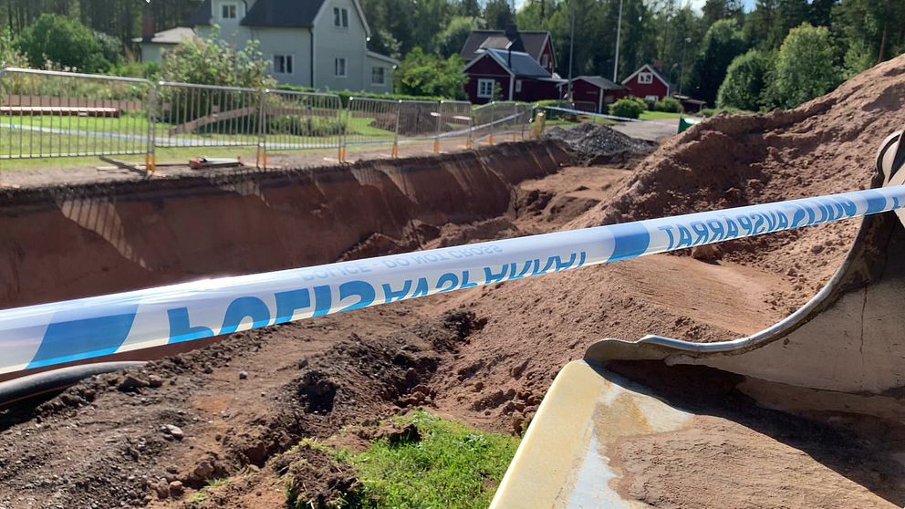 En grävskopa och polisavspärrning vid en grävarbetsplats. Bostadshus i bakgrunden.