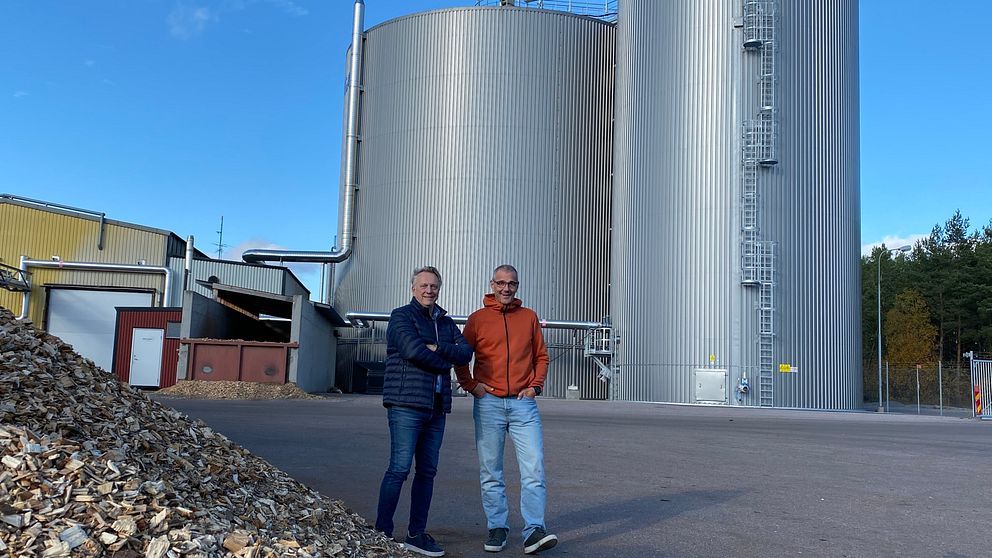 Två personer står framför en industriell anläggning med stora cylindriska tankar i metall. I förgrunden syns en hög med träflis. Bakgrunden består av en klar blå himmel och skog.