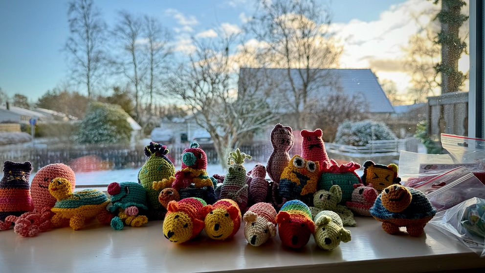 Färgglada, handgjorda virkade djur av olika slag står på en fönsterbrunn med snöiga träd och en klar himmel i bakgrunden.