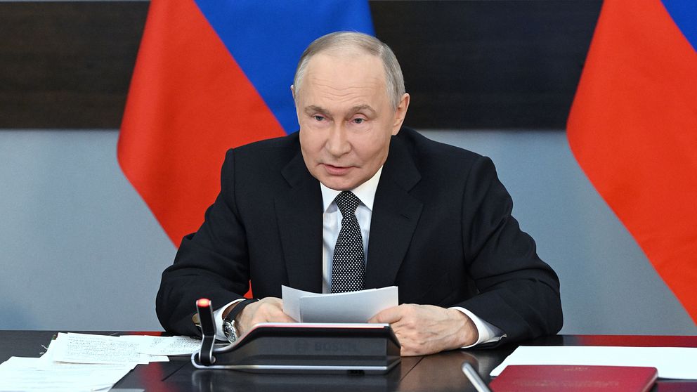 Vladimir Putin i kostym vid skrivbord med ryska flaggor i bakgrunden.