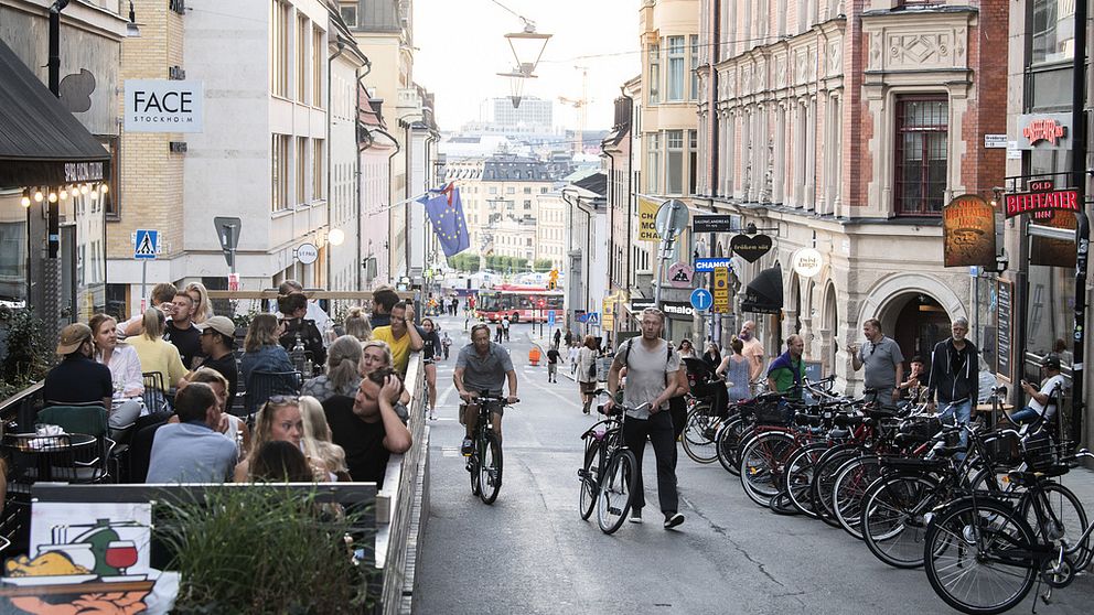 Folk som går och cyklar och fullsatt uteservering i sommarväder på en gata i Stockholm.