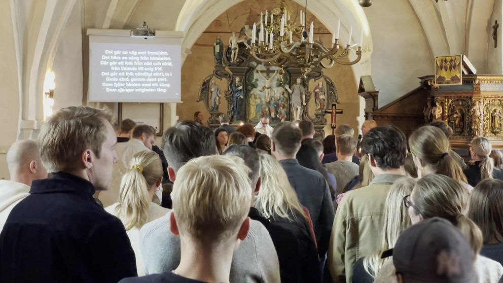 Människor samlas i en kyrka för en ceremoni, med en stor altarvägg och religiösa konstverk i bakgrunden. På en skärm i förgrunden visas text, vilket tyder på en presentation eller hymn. Atmosfären är stillsam och reflekterande.