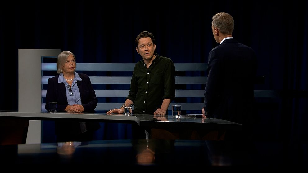 Tre personer står vid ett podium under en studiodiskussion. Det finns glas vatten på bordet och en mörk bakgrund som framhäver scenen.
