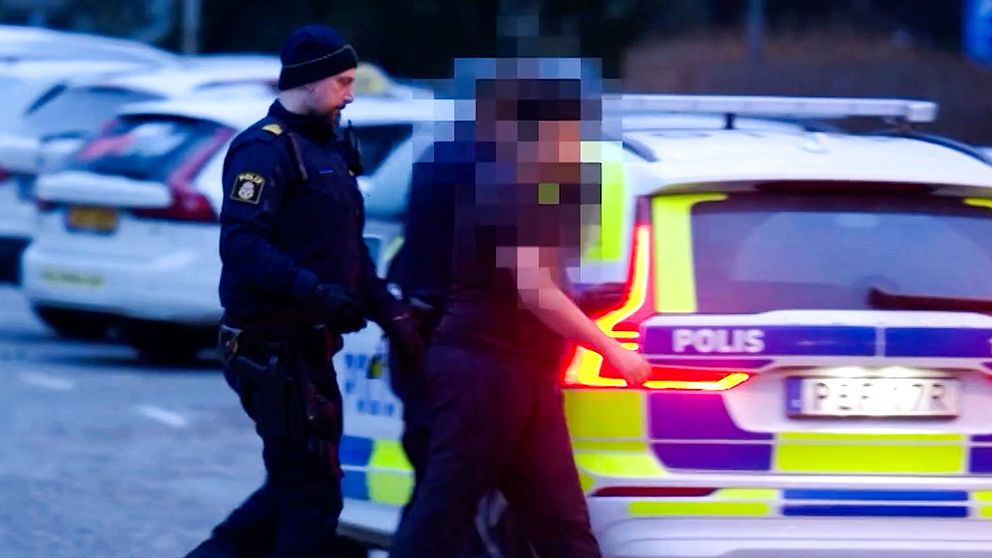 Två poliser eskorterar en person mot en polisiär bil på en parkeringsplats, med andra bilar synliga i bakgrunden.