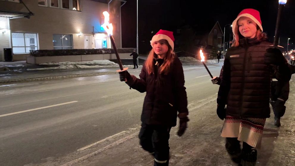 Två personer går längs en gata under natten, bärande på lågor som lyser upp omgivningen. De har på sig tomteluvor och är klädda i varma kläder. Vinterlandskapet har en del snö på marken och byggnader syns i bakgrunden.