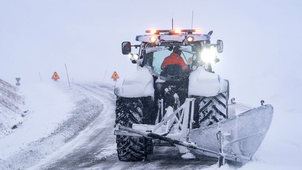 Traktor plogar en snöig landsväg under dimmiga förhållanden.