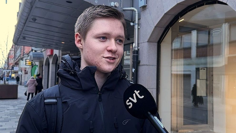 En person står på en trottoar i en stadsmiljö, klädd i en mörk jacka och bär en ryggsäck. En mikrofon med SVT:s logotyp sträcks fram mot personen, som antagligen deltar i en intervju. I bakgrunden syns fler människor, byggnader och en parkerad bil.