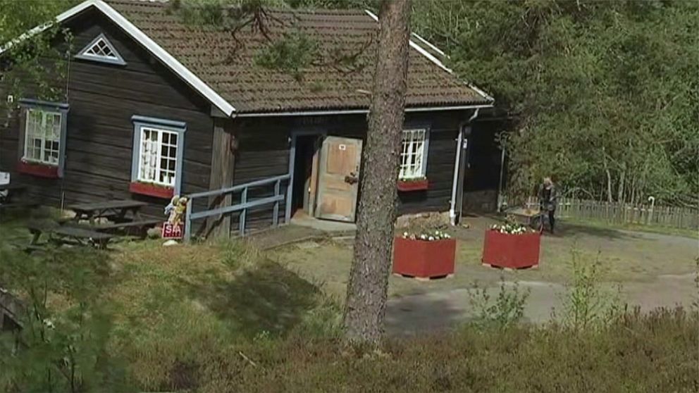 En röd stuga med vita fönster och blomlådor utanför, omgiven av grönskande skog. I förgrunden finns en grusväg och stora blomkrukor. Till höger ses en person röra sig bort från huset.