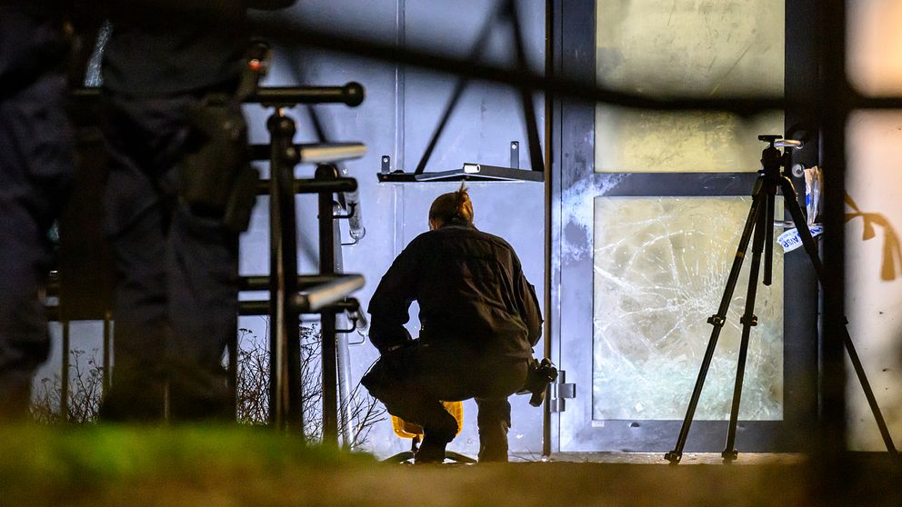 En person i svart klädsel sitter på huk vid en dörr med trasig glasruta. I bakgrunden syns flera poliser samt kameror som är riktade mot platsen. Platsen är ett flerbostadshus på Linero i Lund