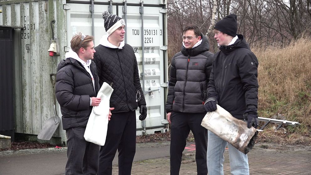 Anton Månsson, Hugo Gårdhagen, Max Albertsson och Anton Johansson Kajrup står bredvid en metallcontainer och håller i två brandsäkra vita påsar. Den ena är ny och den andra är använd och således bränd. Området runt dem är utomhus med viss vegetation. Alla bär vinterkläder.