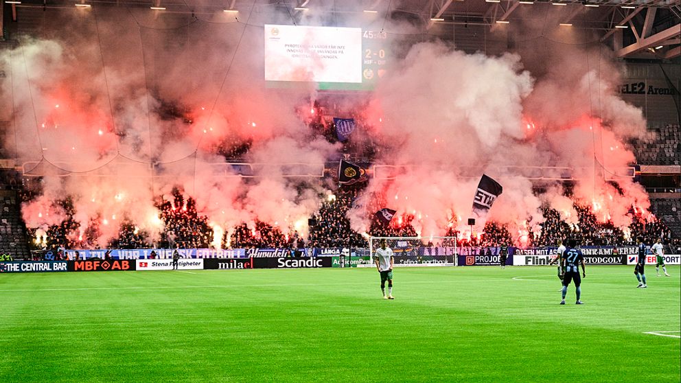 Fulsatt läktare fylld av bengalrök. Spelare ute på en fotbollsplan.