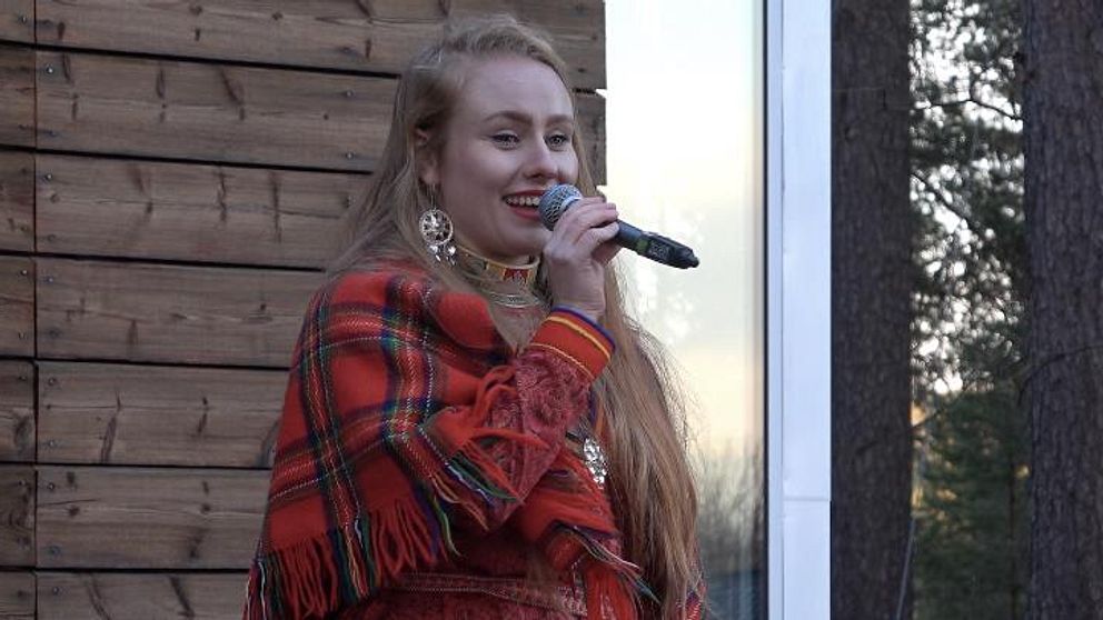 Liv Aira håller en mikrofon iklädd kolt och hälsa alla välkommen till invigningen av jillát