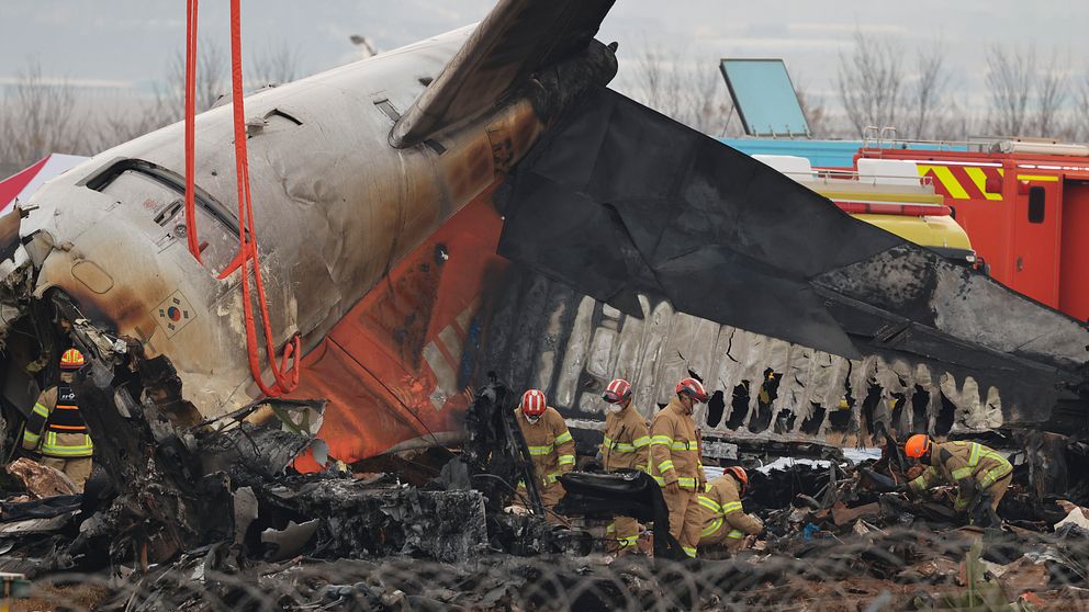 Brandmän arbetar vid vraket av ett kraschat flygplan som har tagit mycket skada efter en brand, med delar av planet delvis förstörda och rök som fortfarande stiger.