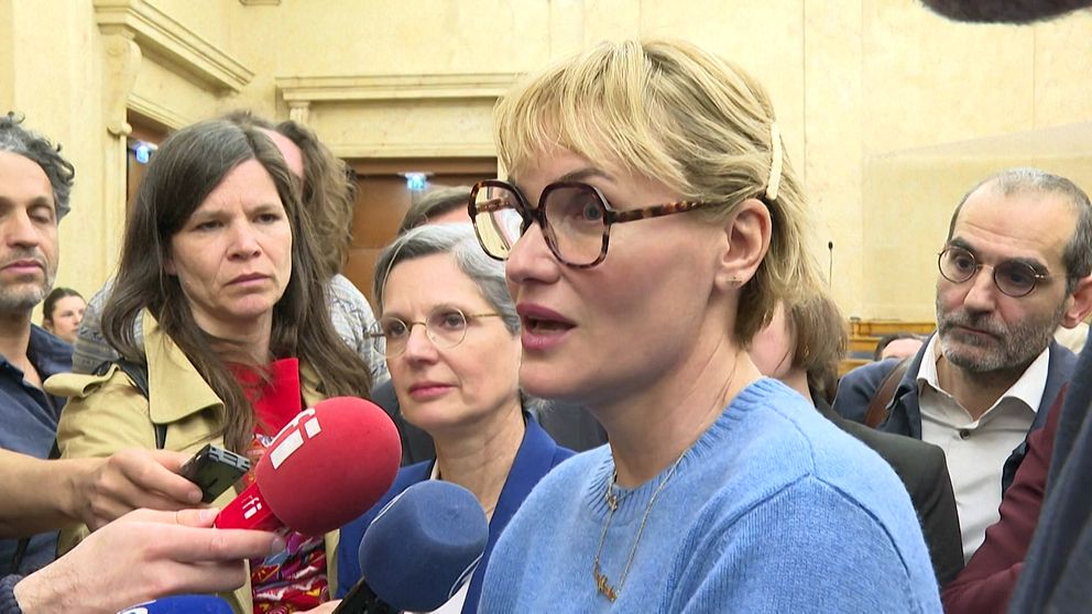 Journalister intervjuar en person inomhus, mikrofoner riktade mot den.