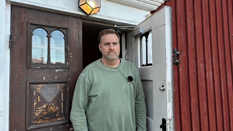 En man står i dörröppningen till en röd stuga. Dörren är öppen och på väggen ovanför dörren hänger en lampa. Bakom mannen syns en del av interiören i stugan.
