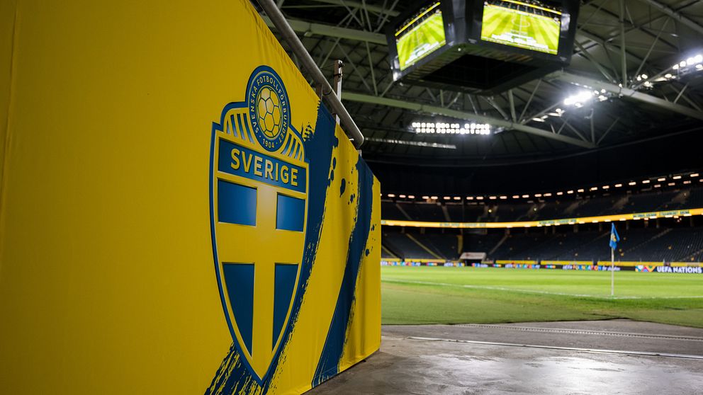Insidan av Friends Arena i Stockholm med det svenska fotbollslandslagets emblem på en gul vägg i förgrunden och en nästan tom fotbollsplan upplyst i bakgrunden.
