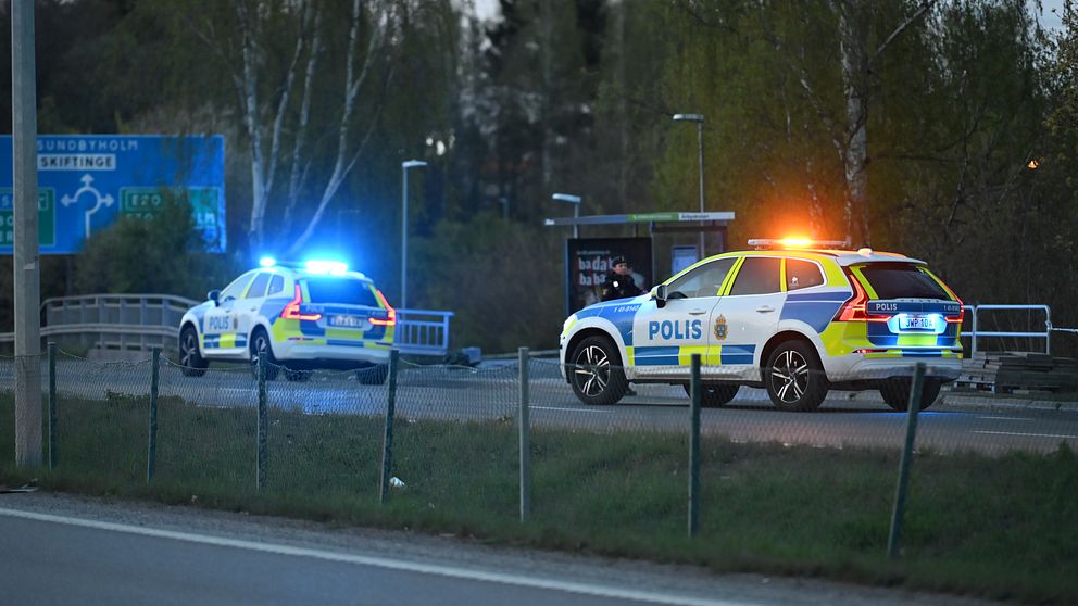 Två svenska polisfordon med blåljus står vid vägkant nära en rondell på kvällen.