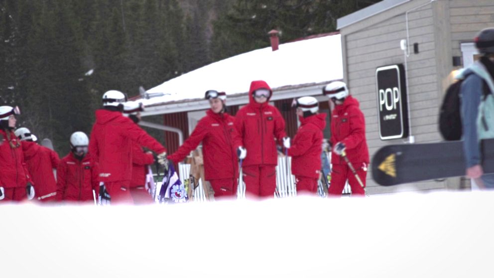 En grupp skidlärare i röda jackor från Skistar står i rad utanför ett hus i Åre. De förbereder sig för att sätta igång med dagen.