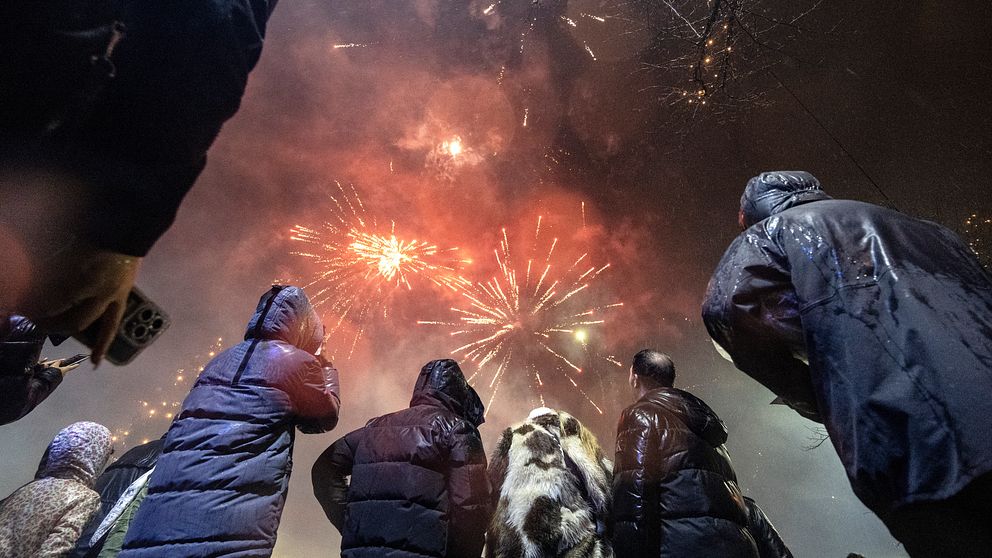 Personer i mörka kläder står i en grupp med ryggen mot kameran och tittar upp på en färgsprakande fyrverkerishow mot en mörk himmel. Fyrverkerierna lyser upp natten med röd, gul och vit färg, medan rök sveper genom scenen.