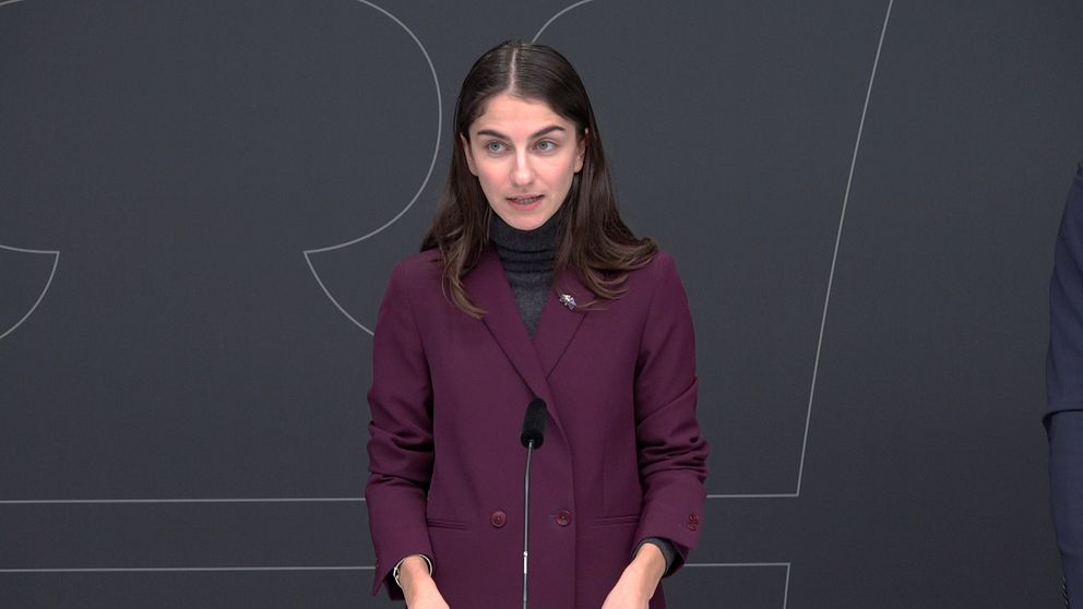 En person står vid ett podium under en presskonferens, iklädd en lila kappa och en mörk tröja. Det finns en mikrofon framför personen, och en mörk bakgrund med grafiska inslag syns bakom.