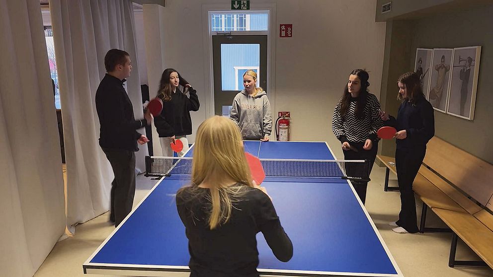 En grupp på fem personer spelar bordtennis i en inomhusmiljö. En person står på ena sidan av bordet och håller en racket, medan de andra fyra står på motsatt sida, beredda att spela. Rummen har ljusa väggar och en stor dörr i bakgrunden. Bänkar syns längs ena väggen.