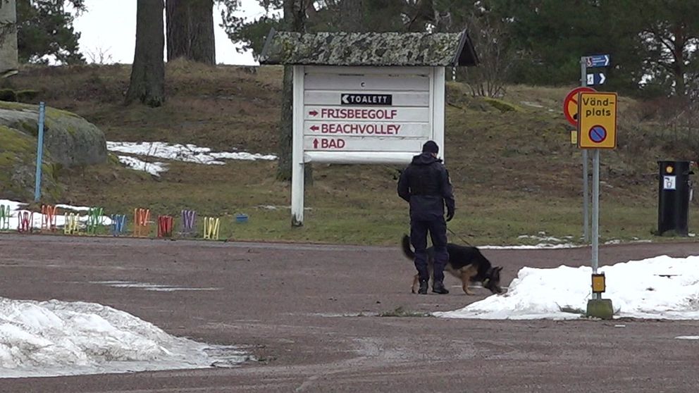 En polis går med en hund på en snötäckt väg bredvid en stor skylt som visar vägbeskrivningar till frisbeegolf, beachvolley och bad på en vintrig dag.