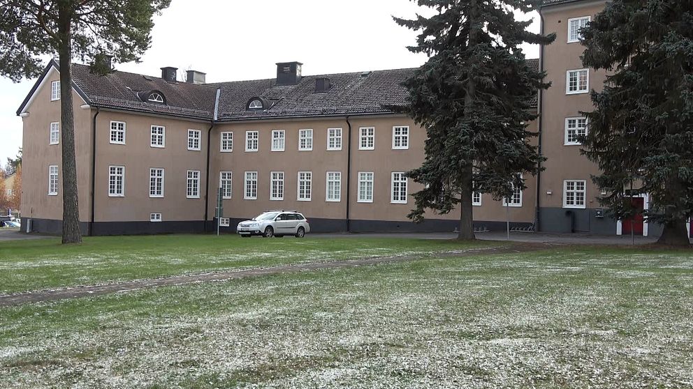 En byggnad av tegel med snö på marken framför, en vit bil står parkerad.