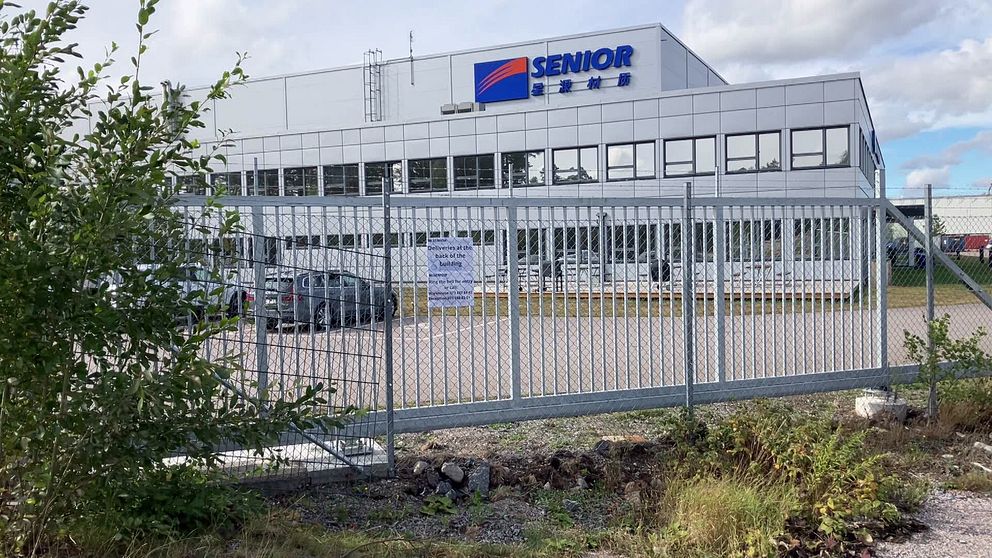 Industribyggnad med skylten SENIOR och parkeringsplats bakom ett metallstängsel.