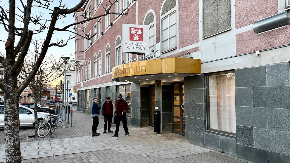 Entrén till Hudiksvalls kommuns byggnad med en skylt som visar kommunens namn. Två personer står vid ingången, med en cykel parkerad i bakgrunden. Fasaden är delvis i grått och rosa, och det är en klar himmel.
