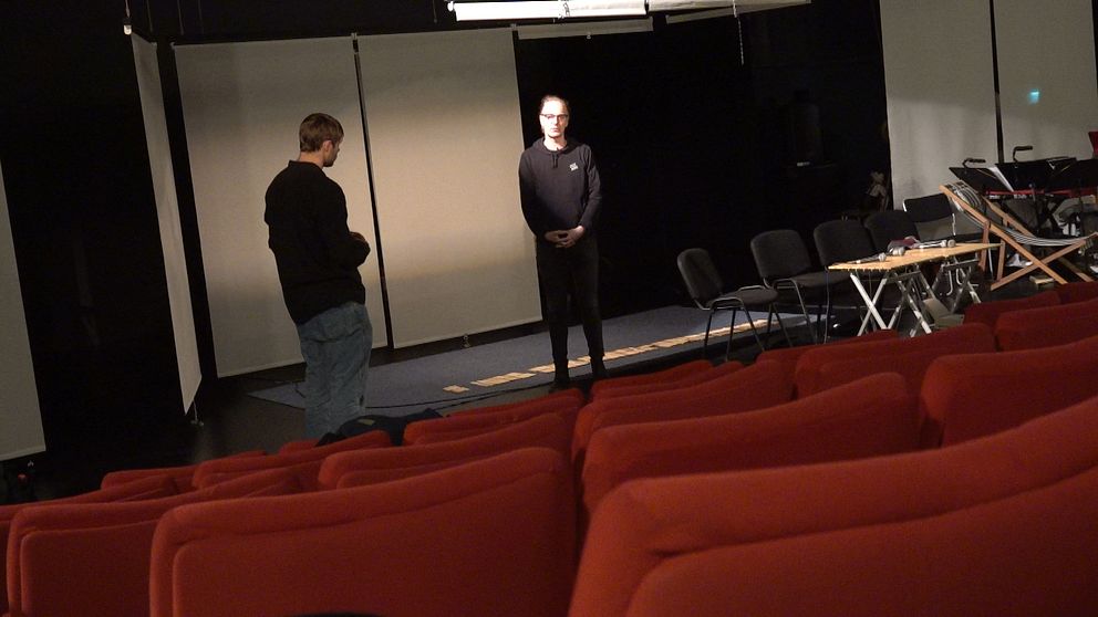 Två män står på en scen, röda stolar i förgrunden.