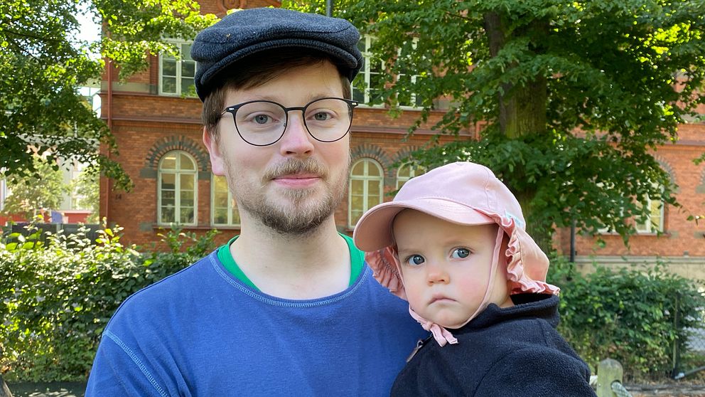 Man med keps och glasögon håller barn med rosa solhatt.