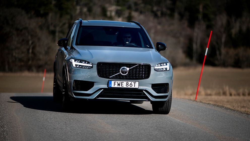 En Volvo XC90 körs på en landsväg med åkrar och skog i bakgrunden, med soligt väder vilket kastar skuggor på vägen. Registreringsskylten visar ”FWE 86T”.