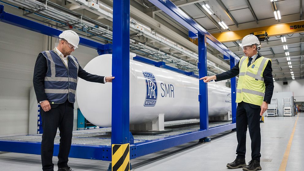 Två personer i skyddshjälmar och västar står vid en vit tank med texten ”SMR”.