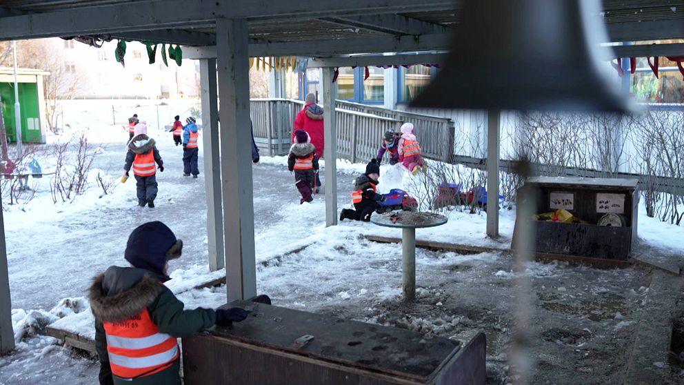Barn i reflexvästar leker utomhus i en snöig förskolemiljö. Några barn springer och leker runt en lekställning, medan andra är sysselsatta vid en bänk. En del av bilden visar en struktur med en rund bordsskiva i förgrunden. Det är en kall och klar dag, och träden är utan löv.