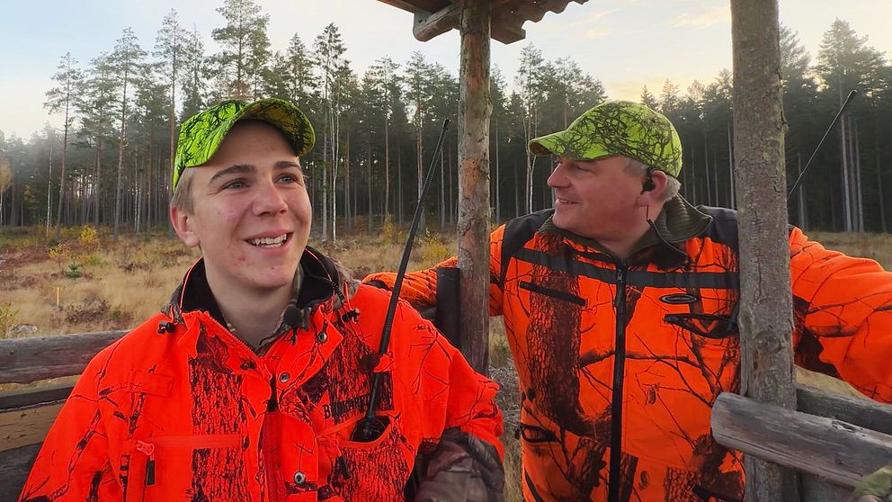 Två personer klädda i orange jaktkläder och matchande kepsar står i ett jakttorn med en skog i bakgrunden. Det är en solig dag, och omgivningen visar höga träd och en öppning i landskapet.