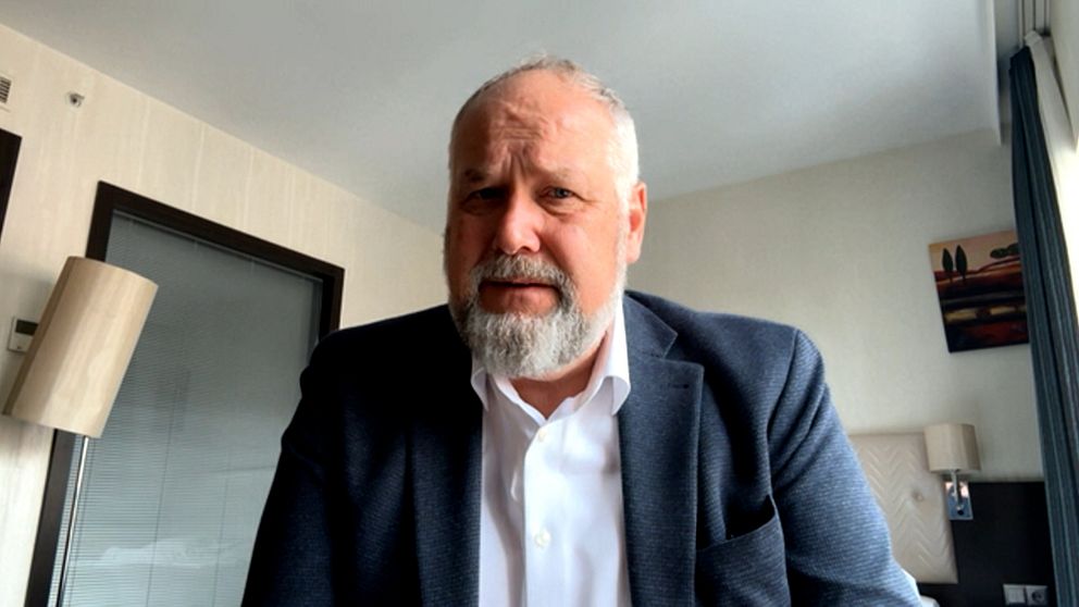 En person i kostym sitter i ett modernt kontorsrum med tavlor och lampa i bakgrunden.