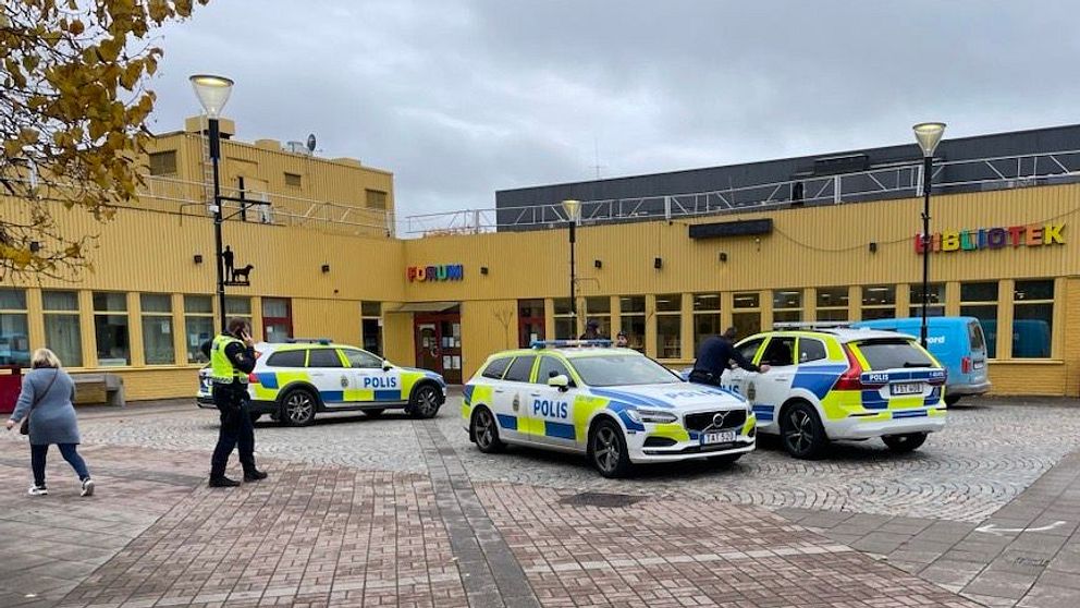 Polisbilar parkerade utanför ett bibliotek, med polispersonal som arbetar på plats. Omgivningen är en torgliknande yta med träd och byggnader i bakgrunden. Det är mulet väder.