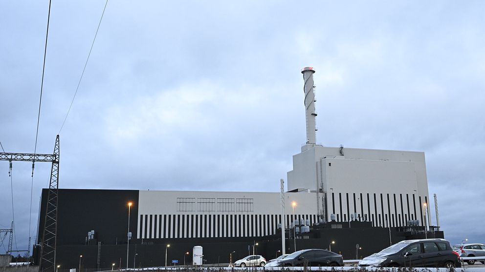 Kärnkraftverket i Oskarshamn. Mörka moln döljer himlen och snö täcker marken. Parkerade bilar syns i förgrunden, medan elledningar sträcker sig över bilden.