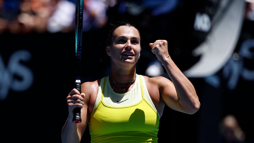 En tennisspelare i en gul tröja håller upp sitt racketslag, även medan hon visar entusiasm med en knuten näve. I bakgrunden syns åskådare som följer matchen.